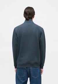 Sweatshirt azul escuro com uma gola alta, mangas longas e punhos canelados. Textura suave. Usado com jeans de ganga azul claro.