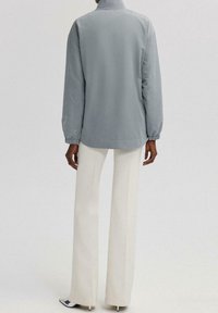 Lys grå jakke med standkrave og elastiske manchetter, med diskrete broderede detaljer på ærmerne. Sæt sammen med hvide bredbenede bukser.