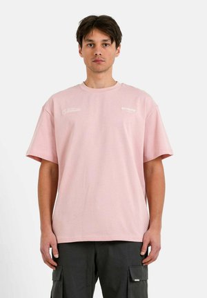 T-shirt en coton rose à manches courtes, col rond, avec des logos blancs sur le devant. Le modèle est associé à un pantalon cargo noir.