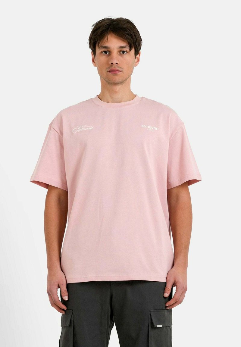 Camiseta de algodón rosa con mangas cortas y cuello redondo, que presenta logotipos blancos en la parte delantera. Modelo combinado con pantalones cargo oscuros.