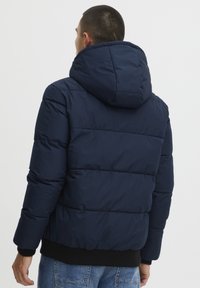Marine blauwe pufferjack met een capuchon, doorgestikte ontwerp en geribbelde manchetten. Het materiaal lijkt glad met een matte afwerking.