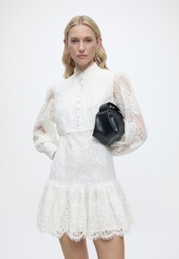 Γυναίκα που φοράει ένα λευκό δαντελένιο φόρεμα με φουσκωτά μανίκια και κουμπιά, κρατώντας μια μικρή μαύρη δερμάτινη τσάντα κάτω από το μπράτσο της.