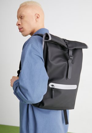 Persoon in blauw shirt draagt een zwarte rugzak met rolltop, voorzien van een voorvak met rits en een gesp, tegen een effen achtergrond.