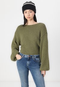 Olivgrön stickad tröja med vida ärmar, kombinerad med blå jeans. Modellen bär en svart mössa med vita ränder.