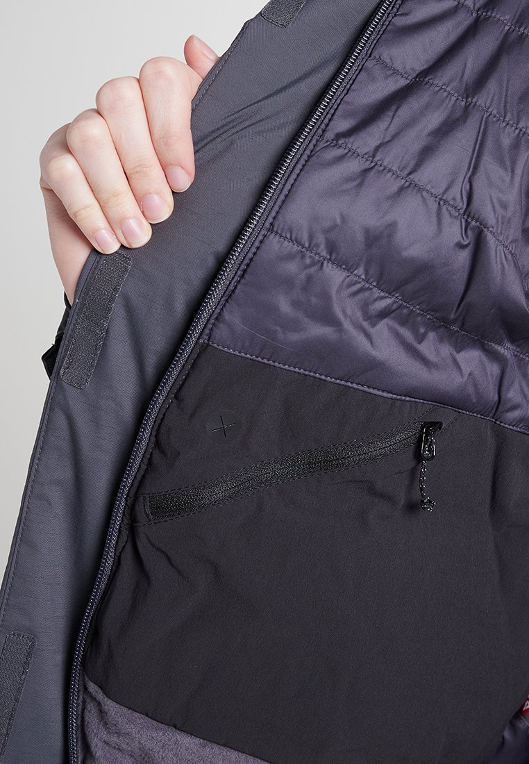 Mano sosteniendo el interior de una chaqueta gris abierta, mostrando un bolsillo negro con cremallera y un forro acolchado con cierres de Velcro.