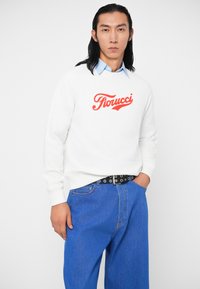 Sweatshirt branca com o logotipo vermelho "Fiorucci", combinado com jeans de ganga azul e um cinto preto com ilhós prateados. Design simples e ajuste casual.