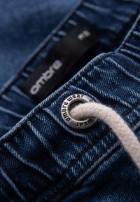Pantalones cortos de mezclilla en azul profundo, con un ojal plateado y un cordón de color crema. Etiqueta con el logotipo visible cerca de la cinturilla.