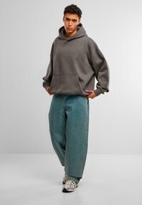 Felpa grigia oversize con tasca frontale, abbinata a jeans azzurri a gamba larga. Il modello indossa sneaker grigie e argentate.