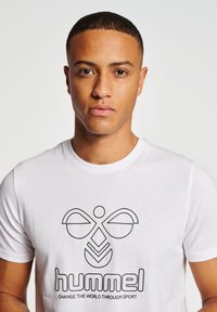 Hvid bomulds t-shirt med et sort grafisk logo design, der indeholder abstrakte former og teksten "hummel" og "FORANDR VERDEN GENNEM SPORT."