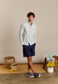 Heren overhemd met korte mouwen heeft een lichte achtergrond met kleurrijke bloemenpatronen. Gecombineerd met marineblauwe shorts en grijze sneakers.