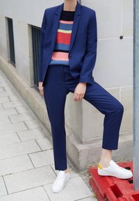 Marinblå kavaj och byxor i kombination med en flerfärgad randig tröja; vita sneakers med gula accenter. Slät tyg, figursydd design.
