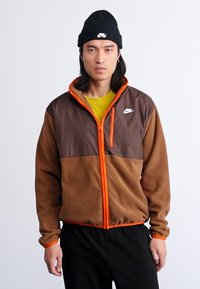 Giacca in fleece marrone Nike con dettagli arancioni, chiusura con zip, colletto alto e logo. Chi la indossa ha anche un berretto nero e una maglietta verde sotto.