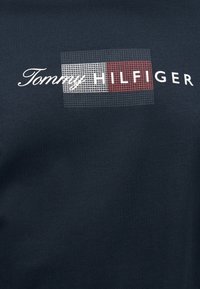 Ύφασμα ναυτικού μπλε με το κείμενο "Tommy Hilfiger"· "Tommy" σε λευκή γραφή με καλλιγραφικά γράμματα, "HILFIGER" με λευκά κεφαλαία γράμματα πάνω σε κόκκινα και λευκά τετράγωνα πλέγματος.
