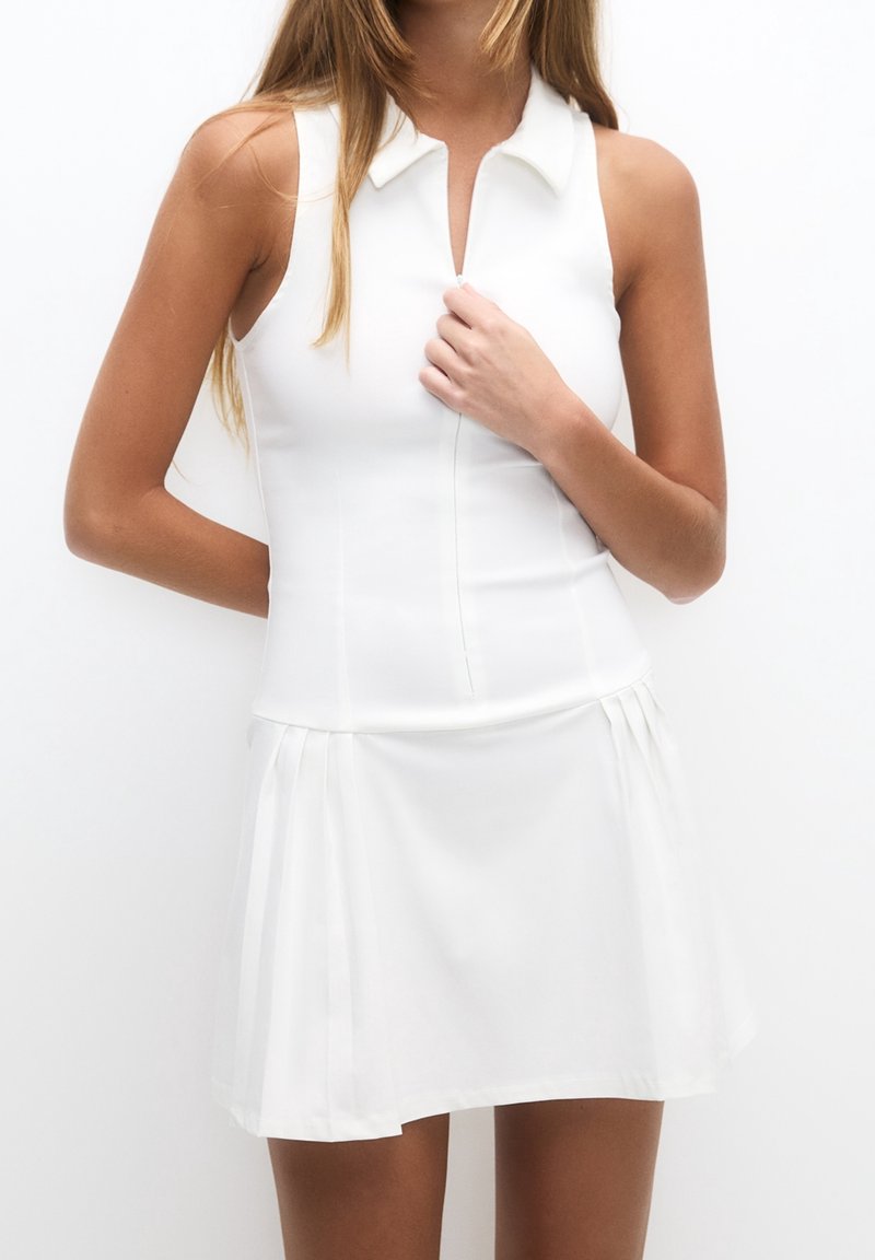 Femme portant une robe blanche sans manches à col avec une fermeture éclair avant et une jupe plissée, debout devant un fond blanc uni.