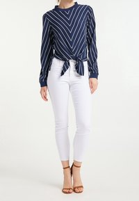 Marineblaue Langarmbluse mit weißen Chevronstreifen und einem geknoteten Front. Getragen mit enganliegenden weißen Jeans und braunen Riemchen-Absätzen.