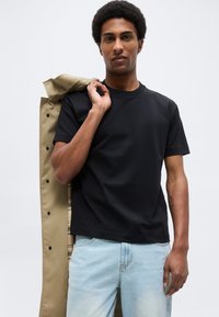 Άνδρας με μαύρο μπλουζάκι και ανοιχτό μπλε τζιν κρατάει μπεζ παλτό στον ώμο, με φόντο μια απλή επιφάνεια.