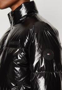Sort pufferjakke lavet af glansmateriale med høj krave, lynlås foran og rund logo-patch på ærmet med røde, hvide og blå accenter.