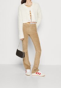 Femme portant un cardigan court en peluche crème, un haut rayé, un pantalon en velours côtelé beige, des baskets blanches avec des rayures rouges, tenant un petit sac à main noir.