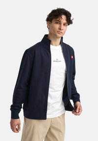 Mørkeblå zip-up hettegenser med høy krage, ribbestrikkede mansjetter og kontrastfarge hvit søm. Brukt over en hvit t-skjorte med logo.