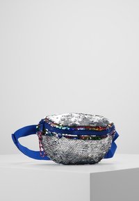 Petite banane argentée à sequins avec fermeture éclair et sangle bleues, présentée sur un socle blanc devant un fond gris clair.