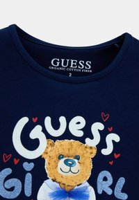 Μπλε ναυτικό οργανικό βαμβακερό t-shirt με σχέδιο καρτούν αρκούδας που φοράει μπλε παπιγιόν και λευκό πουκάμισο, με λαμπερό κείμενο "Guess" και κόκκινες καρδιές ως διακοσμητικά στοιχεία.