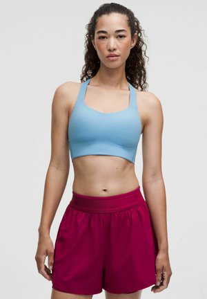 Donna in piedi che indossa un reggiseno sportivo azzurro chiaro e pantaloncini sportivi rosa scuro su uno sfondo grigio chiaro uniforme.