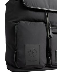 Zaino nero con due tasche anteriori con zip, una delle quali presenta un patch floreale ricamato con la scritta "Ted Baker London."