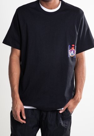 Man draagt een zwart T-shirt met korte mouwen met een kleurrijk Air Jordan-logo op de linkerborst, gecombineerd met zwarte broek.