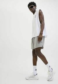 Camisa sin mangas blanca, pantalones cortos gris claro y zapatillas deportivas blancas. El modelo lleva gafas de sol y está en una pose relajada contra un fondo simple.
