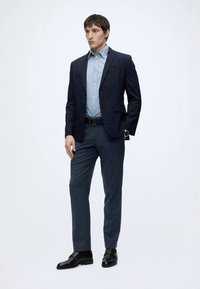 Hombre vestido con chaqueta azul marino, camisa azul clara, pantalones oscuros, cinturón negro, zapatos negros de vestir y reloj de pulsera, de pie frente a un fondo blanco liso.