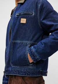 Jachetă din denim de culoare albastru închis, cu guler din catifea, fermoar frontal, buzunar pe piept și buzunare laterale; prezintă cusături vizibile și un etichetă de marcă.