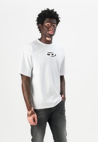 Camiseta blanca de manga corta con un logotipo en forma de corte circular en el pecho. Combinada con jeans gris oscuro, presentada sobre un fondo liso.
