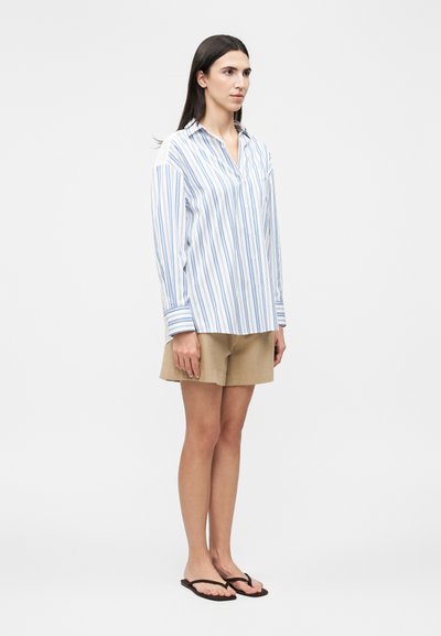 Γυναίκα που στέκεται, φοράει λευκό μακρυμάνικο πουκάμισο με μπλε ρίγες, μπεζ σορτς και μαύρες σαγιονάρες, μπροστά σε λευκό φόντο.