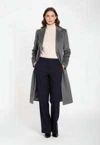 Grauer maßgeschneiderter Mantel mit Spitzrevers und Ein-Knopf-Verschluss, darüber ein beiger Rollkragenpullover. Dunkelblaue Hosen vervollständigen das Outfit.