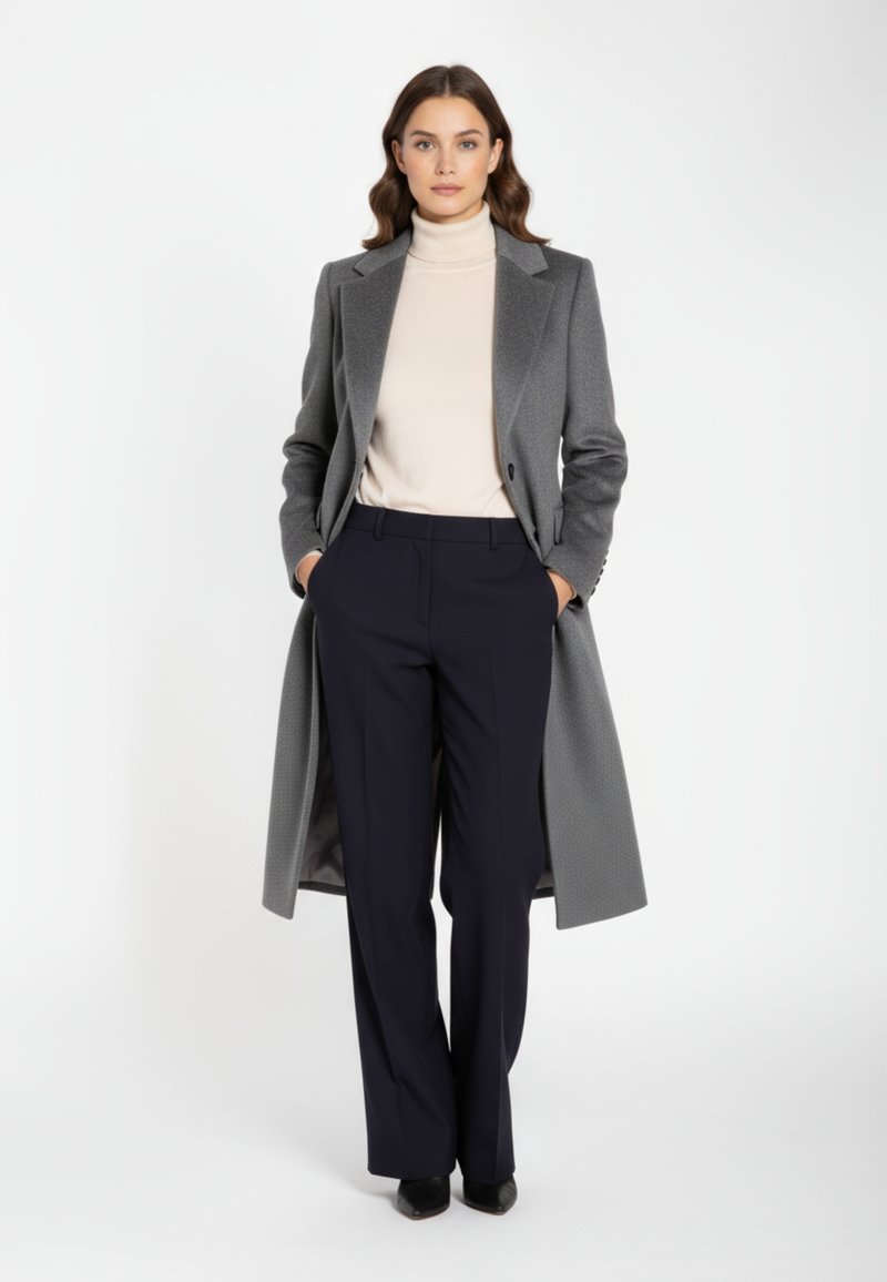 Grauer maßgeschneiderter Mantel mit Spitzrevers und Ein-Knopf-Verschluss, darüber ein beiger Rollkragenpullover. Dunkelblaue Hosen vervollständigen das Outfit.