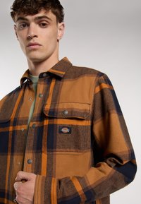 Camicia in flanella a quadri marrone e blu navy con due tasche sul petto e bottoni a scatto in metallo. Presenta un'etichetta tessuta sulla tasca sinistra del petto.