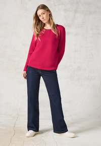Fuchsia stickad tröja med zigzag-mönster och långa ärmar, tillsammans med mörkblå utsvängda jeans och vita sneakers, vilket skapar en avslappnad outfit.