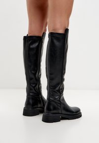 Botas altas de imitación de cuero negras con un acabado suave, punta redondeada y suela texturizada. Presentan una cremallera lateral para facilitar su uso.