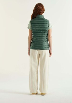 Veste matelassée verte avec des coutures horizontales, col haut et un matériau léger, associée à un pantalon large crème et des chaussures beiges.