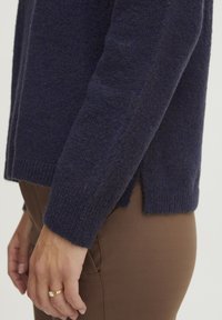 Marineblå strikket sweater med en tekstureret finish. Har en afslappet pasform, sideslids og ribbede ærmer, parret med brune, tætsiddende bukser.
