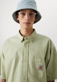 Lichtgroen, gestructureerd shirt met korte mouwen en een borstzak, met Carhartt-logo patch. Het model draagt een lichtblauwe bucket hat en heldere brillen.