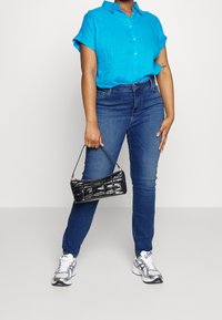 Camicetta turchese a maniche corte, jeans blu, mini borsa nera in vernice con manico e scarpe da ginnastica argento con accenti blu.