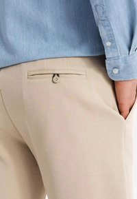 Pantaloni beige con finitura texurizzata, dotati di una tasca posteriore con chiusura a bottone, abbinati a una camicia azzurro chiaro visibile nella parte superiore.