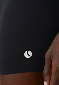 Juodi sportiniai šortai, pagaminti iš lygaus audinio, su balta logo apatinėje dalyje, pritaikyti ir be siūlių kraštu.