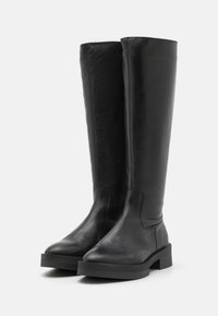 Botas de cuero negro hasta la rodilla con una textura suave, punta redondeada y una gruesa suela de goma negra, que presentan un detalle de costura mínimo.