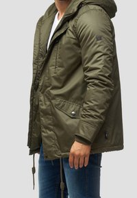 Oliwkowa kurtka ocieplana z kapturem, z przednim zamkiem, dwoma bocznymi kieszeniami oraz regulowanymi sznurkami u dołu. Gładkie, teksturowane wykończenie.