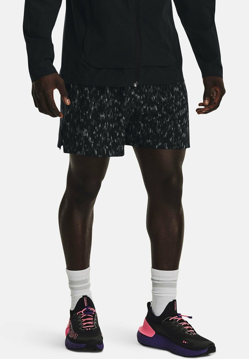 Man med svarta mönstrade träningsshorts, svart jacka, vita strumpor och svarta löparskor med rosa och lila detaljer.