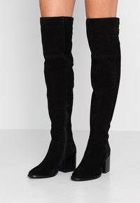 Botas de ante negro hasta el muslo con un tacón grueso, cierre lateral y textura suave. Diseño de punta redondeada y silueta ajustada.