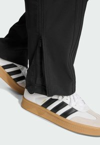 Svarta träningbyxor med dragkedjor på sidorna och vita Adidas Samba-sneakers med svarta ränder och gummisula.