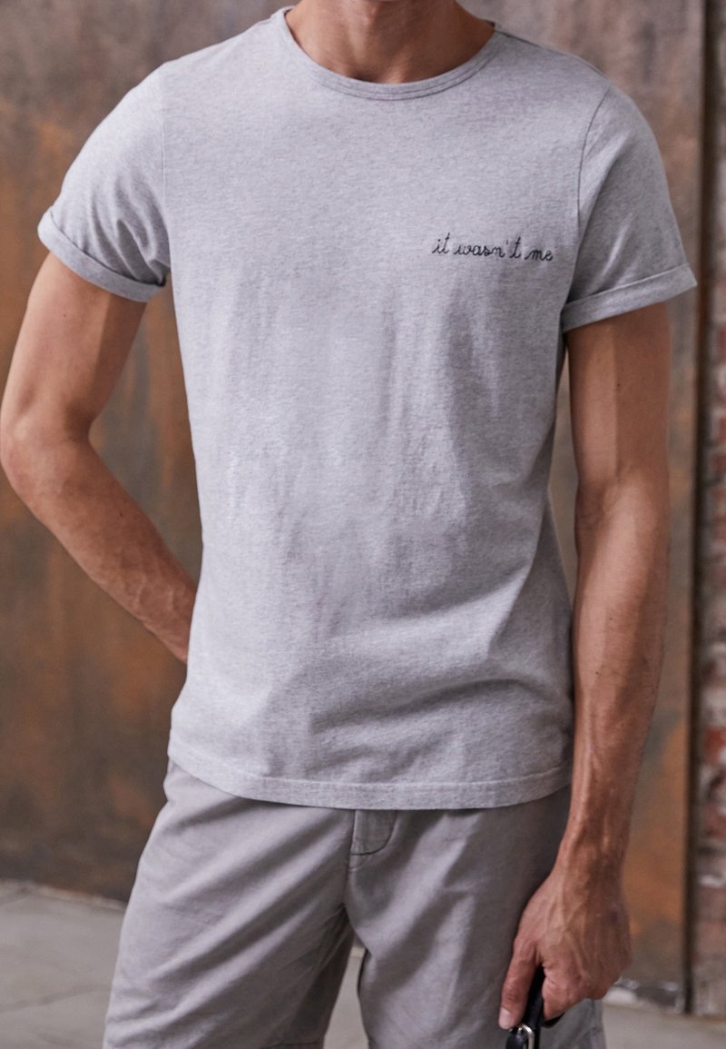 T-shirt gris à col rond avec des manches courtes retroussées, présentant un texte brodé "ce n'était pas moi" sur le côté gauche de la poitrine. Fabriqué en tissu de coton doux.